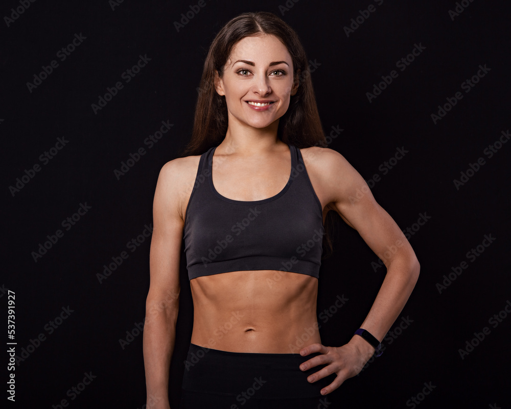 Confident sportswoman in grey sportsbra and shirt, holding hands on waist, fitness trainer standing in power pose, workout in gym isolated on black background