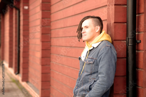 Portrait of young positive man with dreadlocks standing, leaning on wall, on city street.