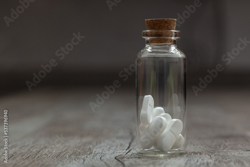 Glass container with white pills on a wooden table. the Container is located to the left of the center picture on a grey background. 
