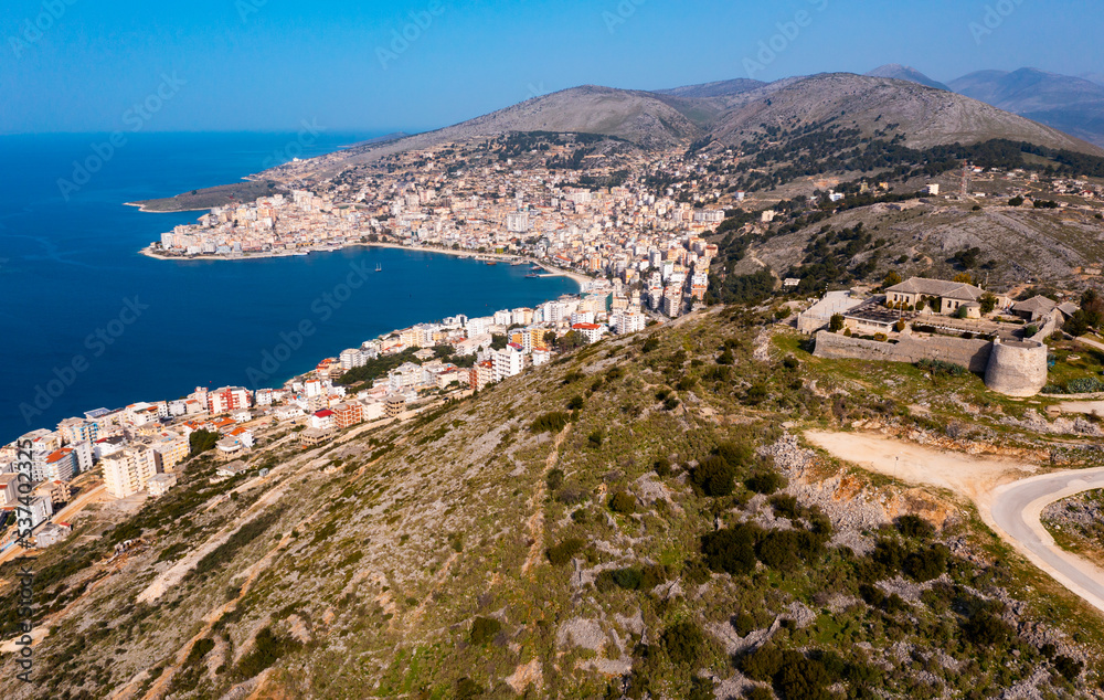 Scenic aerial view of Sarande cityscape on shores of gulf of Ionian Sea and ancient Lekuresi Castle on hill above city on sunny spring day, Albania