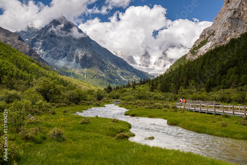 Landscape of summer at Chongu pasture and two tourists walking on the wooden walkway in Yading national level reserve, Daocheng, Sichuan Province, China.