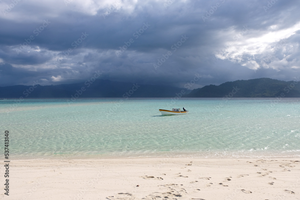 A small motorboat moored in the crystal clear turquoise ocean water of a remote sandbar island, rainstorm and dark clouds in distance, tropics of Bougainville, Papua New Guinea