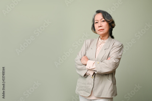 Professional aged-asian businesswoman in casual suit arms crossed, isolated over green background.