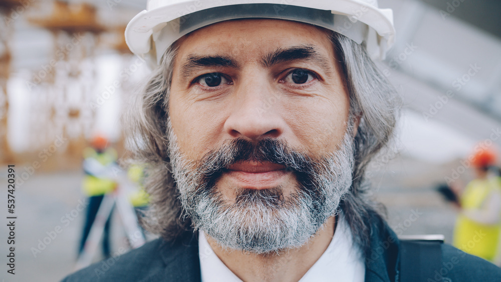 Close-upportrait of mature businessman standing at industrial site wearing helmet and looking at camera. Investors and building concept.