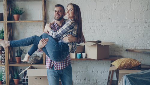 Fototapeta Naklejka Na Ścianę i Meble -  Cheerful strong guy is whirling his girlfriend while moving to new house after purchasing it. Romantic relationship, people and housing concept.