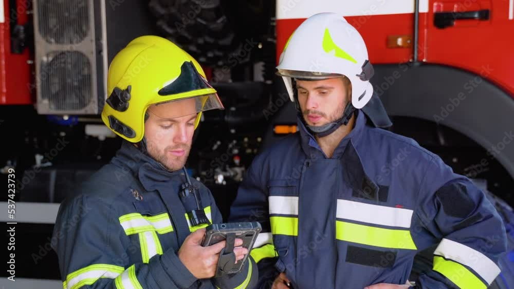 Two firemen in full equipped clothes standing outside at the fire truck with a tablet in hands and deciding what to do for extinguishing fire Two firemen in full equipped clothes standing outside at the fire truck with a tablet in hands and deciding what to do for extinguishing fire