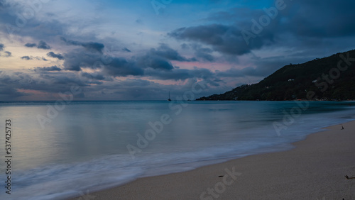 A quiet evening on a tropical beach. The waves of the turquoise ocean are foaming on the sand. Clouds in the sky, highlighted in pink. A hill in the distance. Reflection on the water. Long exposure.