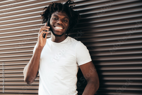 Handsome hipster model.Unshaven African man dressed in white summer t-shirt. Fashion male with dreadlocks hairstyle. Posing in the street. Using his smartphone. Talking with friends at cellphone