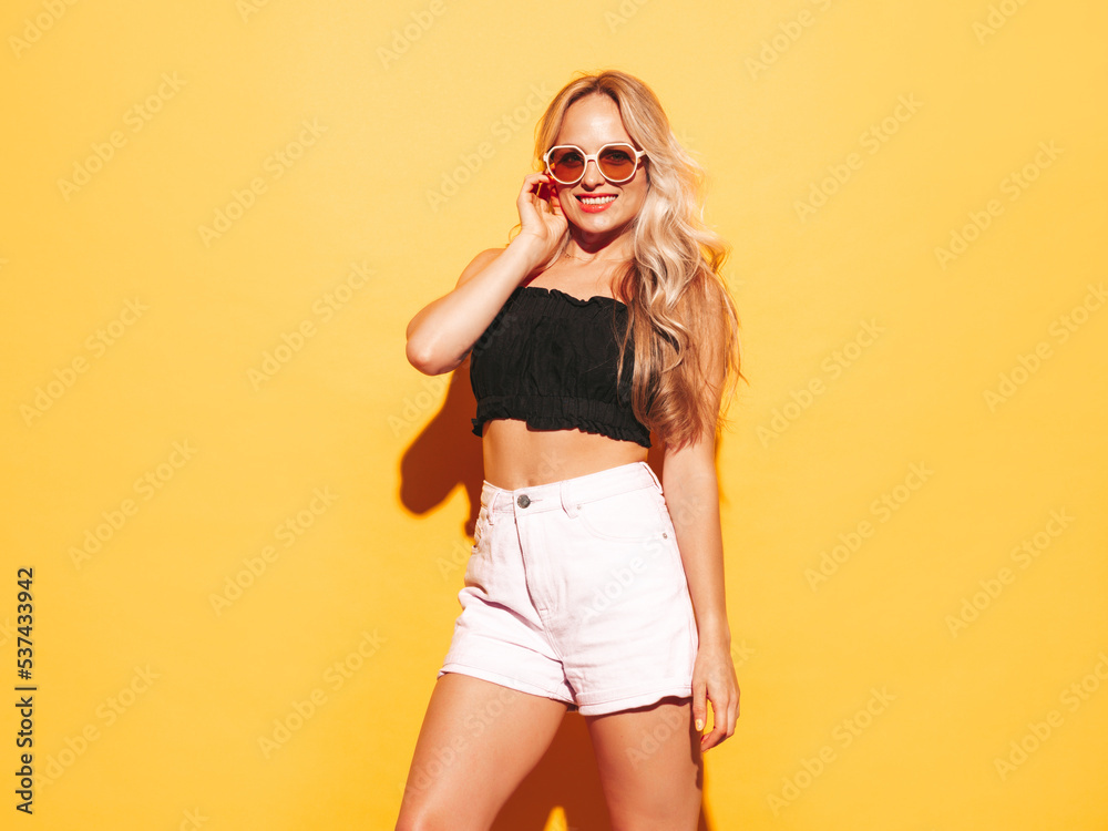 Fototapeta premium Young beautiful smiling blond female in trendy summer shorts clothes. Sexy carefree woman posing near yellow wall in studio. Positive model having fun indoors. Cheerful and happy. In sunglasses