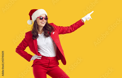 Cheerful joyful confident pretty Santa woman in disco party outfit, Xmas cap, white gloves and sun glasses standing and pointing to side isolated on yellow background. Christmas, New Year concept