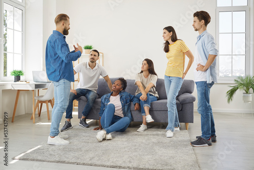 Friends meeting at home. Young people gather in living room, talk, listen to each other, discuss different things. Group of diverse friends listening to man telling story and explaining what happened