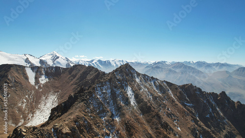 Snowy rocky mountain peaks in a light haze. Aerial view from the drone of the blue sky, light haze and steep cliffs with peaks. A moraine lake can be seen in the distance. Ancient glaciers. Kazakhstan