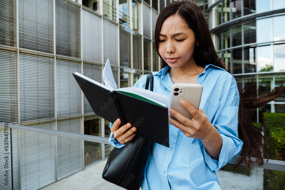 Asian brunette woman using mobile phone while standing with planner Asian brunette woman using mobile phone while standing with planner