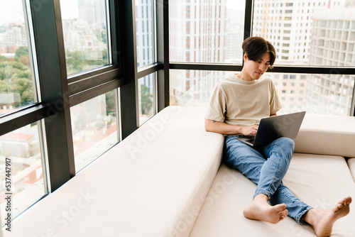 Asian young man using laptop computer while sitting by window at home