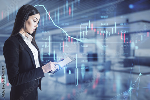 Attractive young european businesswoman using tablet with glowing candlestick forex chart hologram on blurry office interior background. Stock, investment, market and money concept. Double exposure.