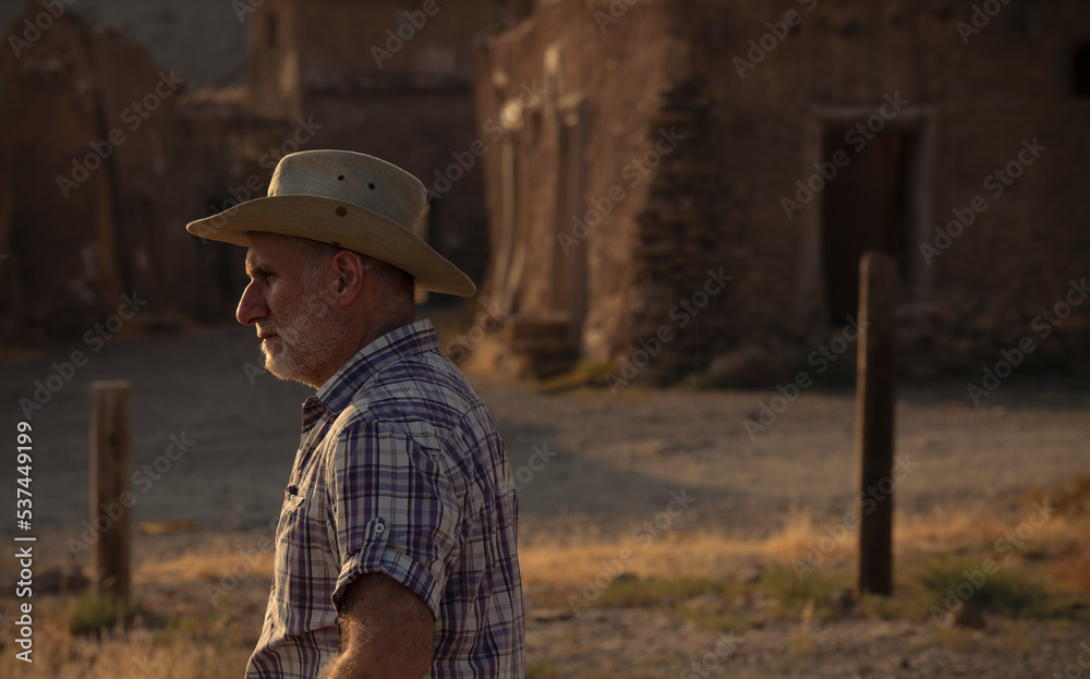 Side view of adult man in cowboy hat and shirt against abandoned building