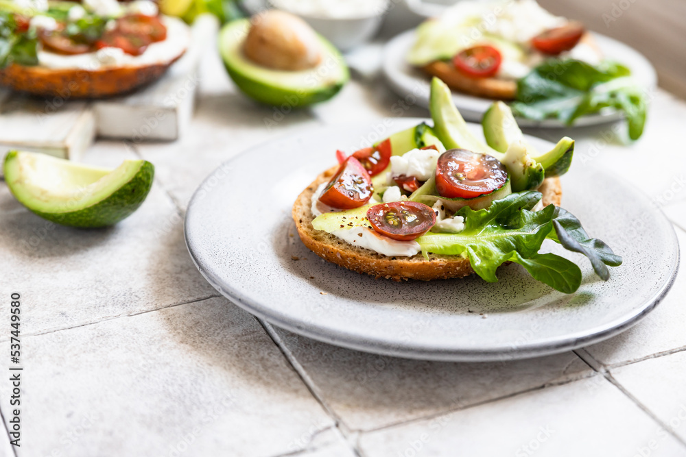 Bagels sandwich topped with cream cheese, farm cottage cheese, avocado, tomatoes, cucumbers and salad, grey tilled background. Healthy breakfast food.