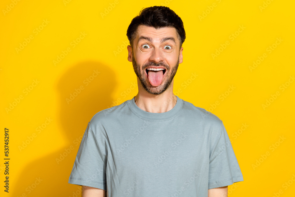 Portrait photo of young funky man bristle brunet stick out tongue positive grimace face crazy good mood weekend isolated on yellow color background