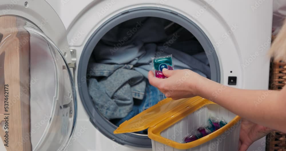 The woman sits at the washing machine and performs household chores. The clothes placed in the drum are waiting for the program to start. The girl holds a box of capsules drops one inside the laundry. The woman sits at the washing machine and performs household chores. The clothes placed in the drum are waiting for the program to start. The girl holds a box of capsules drops one inside the laundry.