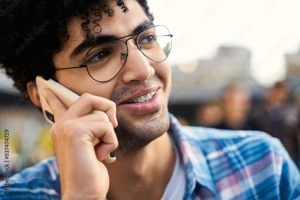 Handsome middle eastern male student calling to friend sitting in cafe. Smiling multiracial hipster guy talking mobile phone outdoors