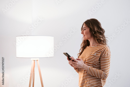 Young woman holding mobile phone beside floor lamp at home photo