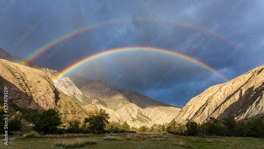 Bright full rainbow on the background of storm cloud over mountain range and valley.