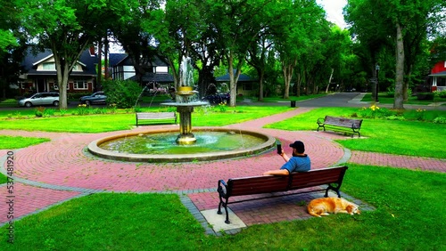 bearded guy black baseball cap looking at phone on a park bench with his golden dog by cobblestone green grass with a post modern metallic fountain sprinkling water at a circle with vintage mansions photo