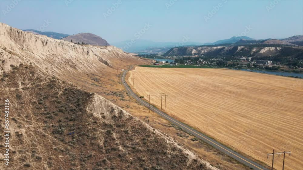 Aerial view of road between mountain and field with hills in the distance. Beautiful drone footage of a mountainous landscape under bright blue sky
