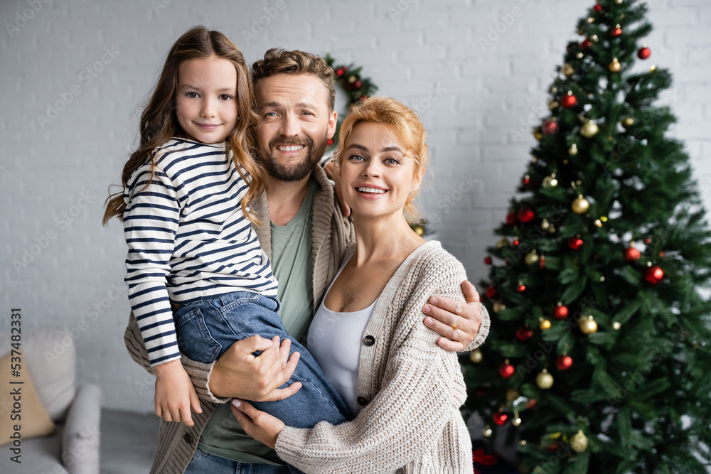 Smiling man hugging wife and daughter near blurred christmas tree at home Smiling man hugging wife and daughter near blurred christmas tree at home