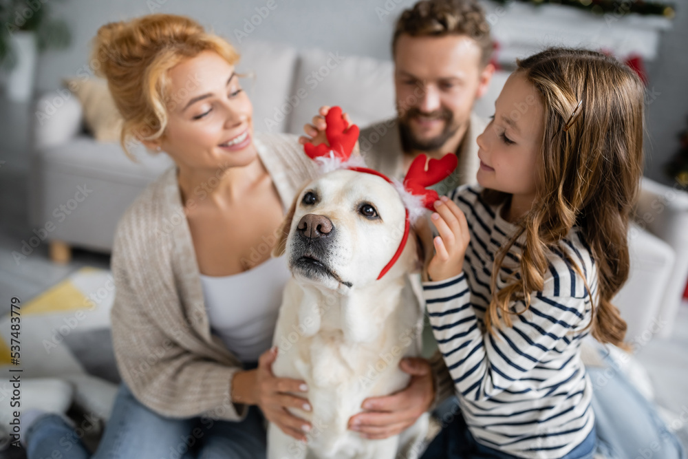 Kid wearing christmas headband on labrador near blurred parents at home