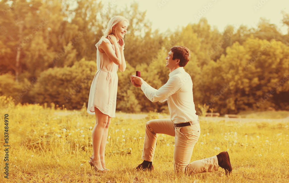 Fototapeta premium Happy young couple, man kneeling down and proposing a ring to his beloved woman outdoors in the park, wedding concept