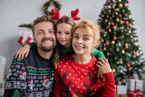Smiling family in christmas sweaters hugging and looking at camera at home