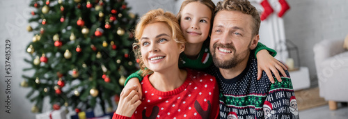 Kid hugging smiling parents in warm sweaters during christmas celebration at home, banner
