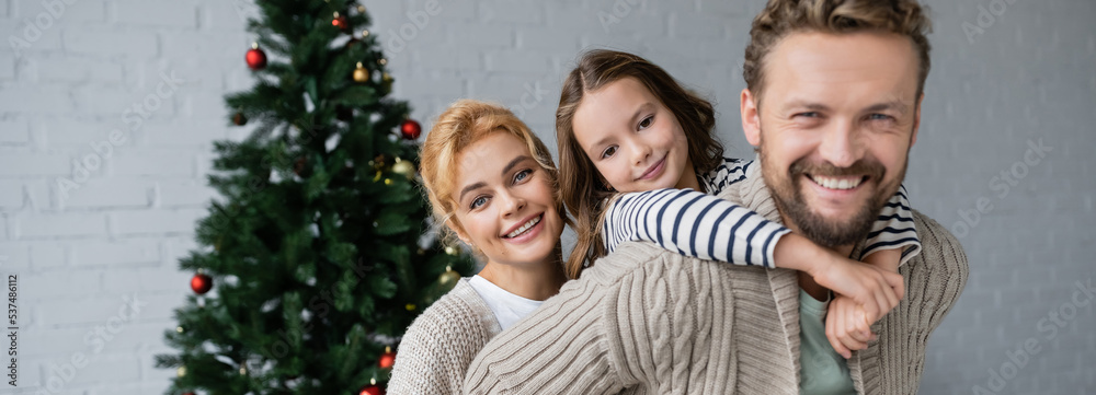 Positive family in warm cardigans looking at camera near christmas tree at home, banner