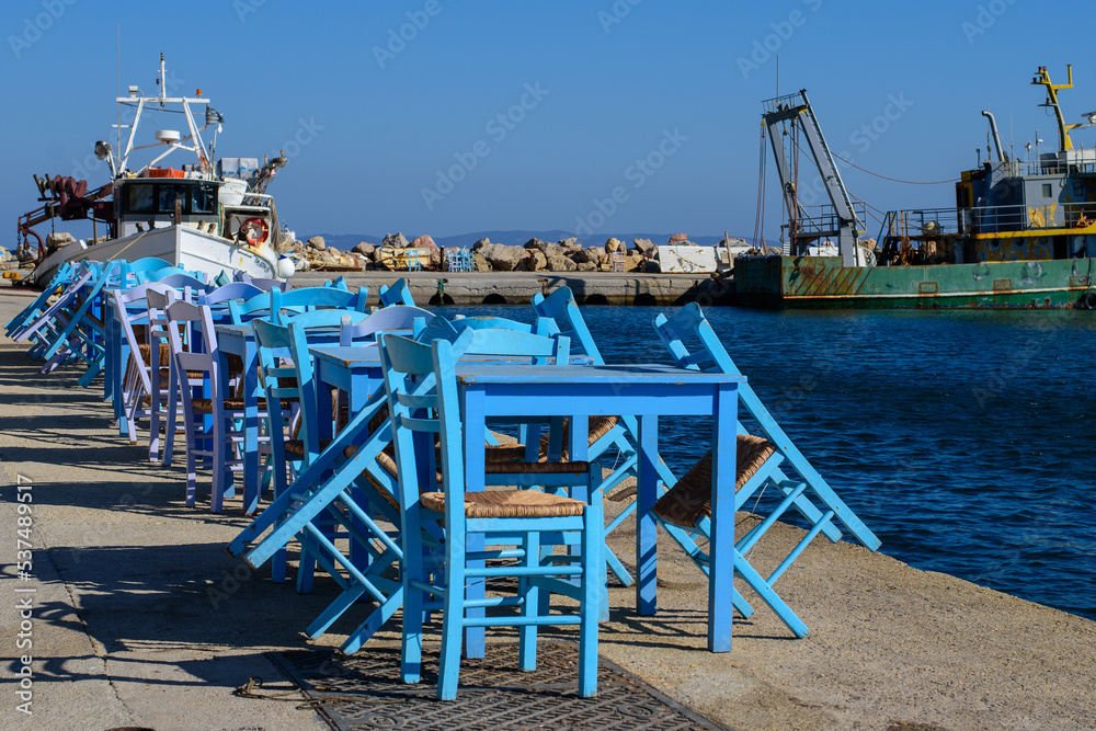 Blue wooden tables and chairs of a typical fish tavern at the port. Fishing ships on the background. Aegean island Chios in Greece on an autumn day. Greek holidays and destinations