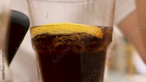 Cold soda cola beverage with lemon slice and ice cubes. Close up shot of a man in white short. drinking coke and staring at the phone in the sunny summer day at a restaurant. Dark soft drink cocktail (ID: 537491399)