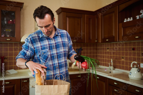 Dark-haired European man in blue checkered shirt, lays out purchases in the kitchen, sorting vegetables and greens while unpacking grocery shopping bag after shopping for groceries