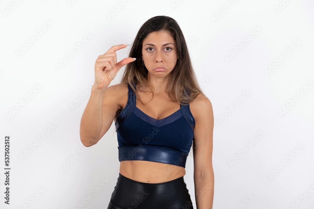 young beautiful woman wearing sportswear over white background purses lip and gestures with hand, shows something very little. young beautiful woman wearing sportswear over white background purses lip and gestures with hand, shows something very little.
