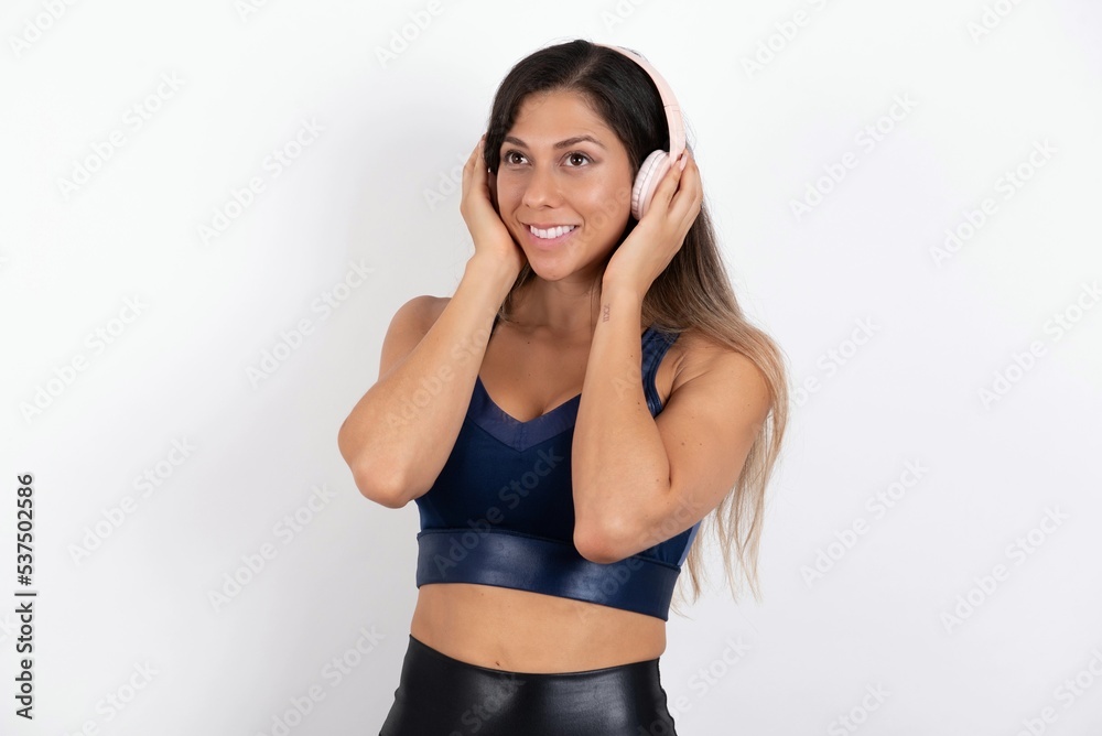 young beautiful woman wearing sportswear over white background wears stereo headphones listens music concentrated aside. People hobby lifestyle concept young beautiful woman wearing sportswear over white background wears stereo headphones listens music concentrated aside. People hobby lifestyle concept
