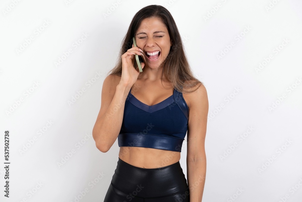 Overemotive happy young beautiful woman wearing sportswear over white background laughs out positively hears funny story from friend during telephone conversation