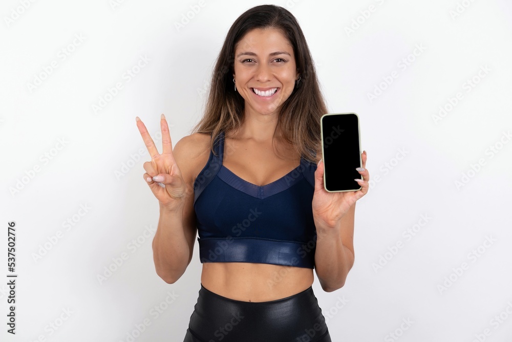 young beautiful woman wearing sportswear over white background holding modern device showing v-sign