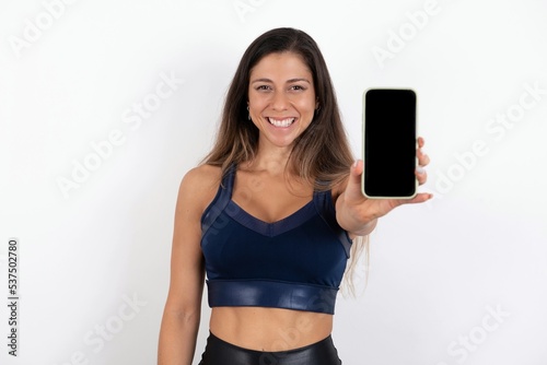 Charming adorable young beautiful woman wearing sportswear over white background holding modern device, showing black screen smartphone