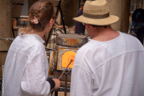 Pareja de espaldas vestidos de blanco elaborando objetos de vidrio soplado de manera tradicional y artesanal en un horno candente durante un mercado medieval en Hondarribia País Vasco