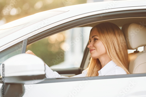 Sitting and driving. Young woman in white clothes is with her electric car at daytime