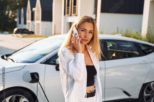 Holding phone and talking by it. Young woman in white clothes is with her electric car at daytime © standret