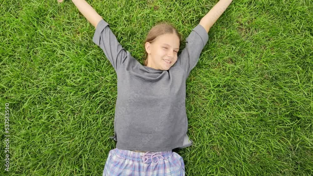 Joyful teen girl lies on grass and wave her hands imitating flapping of a bird wings. Happy child enjoy warm summer day relax on young green grass. Wonderful mood of teenager. Adolescent psychology Joyful teen girl lies on grass and wave her hands imitating flapping of a bird wings. Happy child enjoy warm summer day relax on young green grass. Wonderful mood of teenager. Adolescent psychology