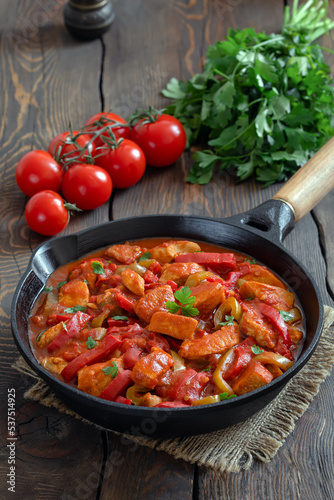 Turkish stew Tavuk sote. Sauteed chicken breast in tomato sauce mixed with mixed with onion and colorful bell pepper served in in frying pan. Wooden background, vertical, selective focus.