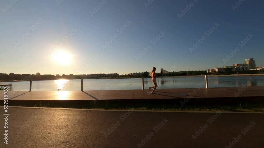 Side FPV drone view of sporty young woman jogging along wooden pier along the city waterfront on bright sunny summer morning. Aerial view of athletic female running on beach by sea, slow motion. Side FPV drone view of sporty young woman jogging along wooden pier along the city waterfront on bright sunny summer morning. Aerial view of athletic female running on beach by sea, slow motion.