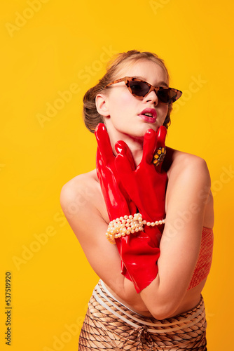 High fashion. Half-length portrait of young fashionable girl in red huge latex glove and sunglasses isolated over bright yellow background. Weird people concept. photo