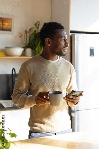 Happy african american man standing in kitchen, drinking coffee and using smartphone